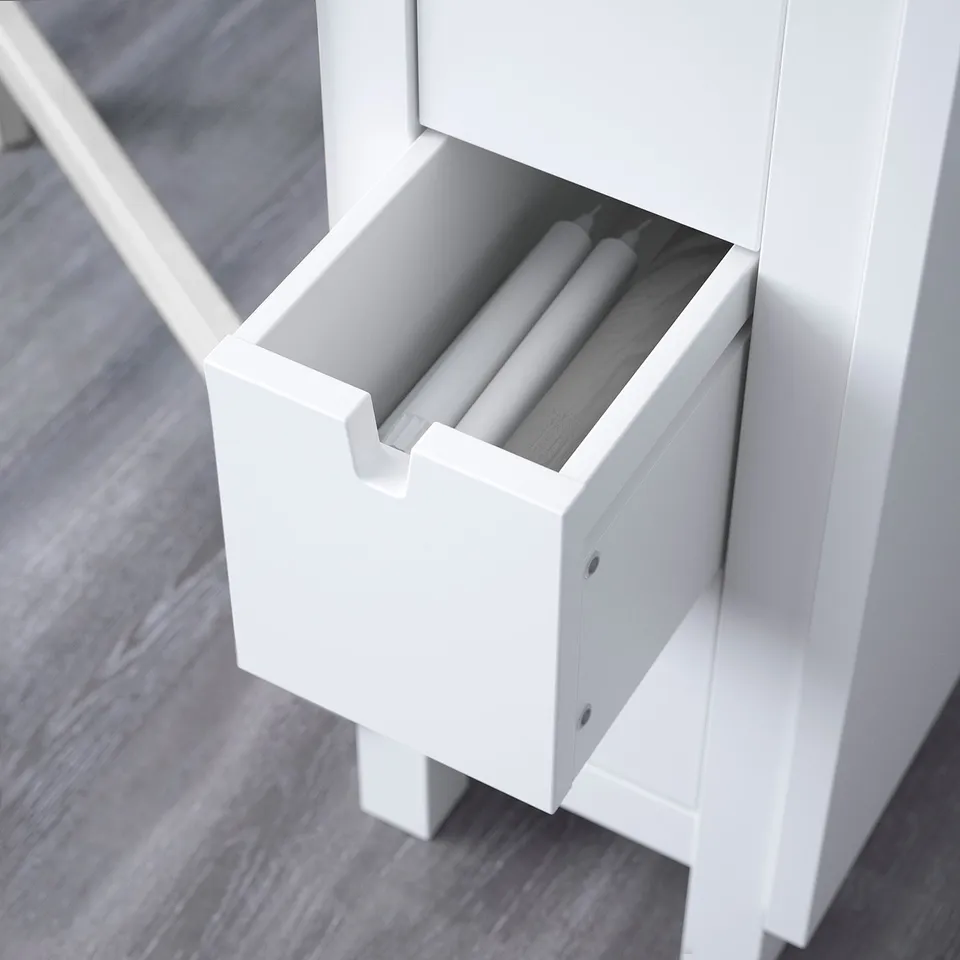 White drawer unit, part of a table set, designed for storing items like cutlery. Open drawer displaying cylindrical items.