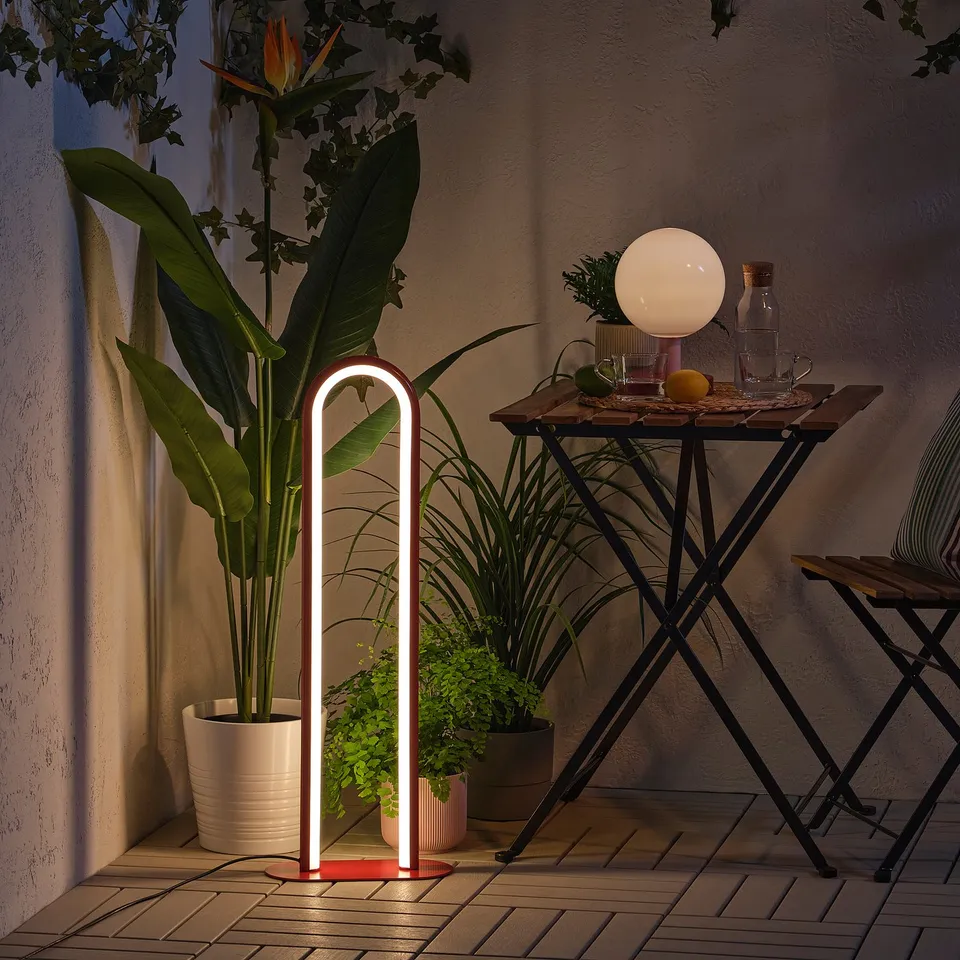 Modern outdoor/indoor lamp, glowing white, stands beside a potted plant.