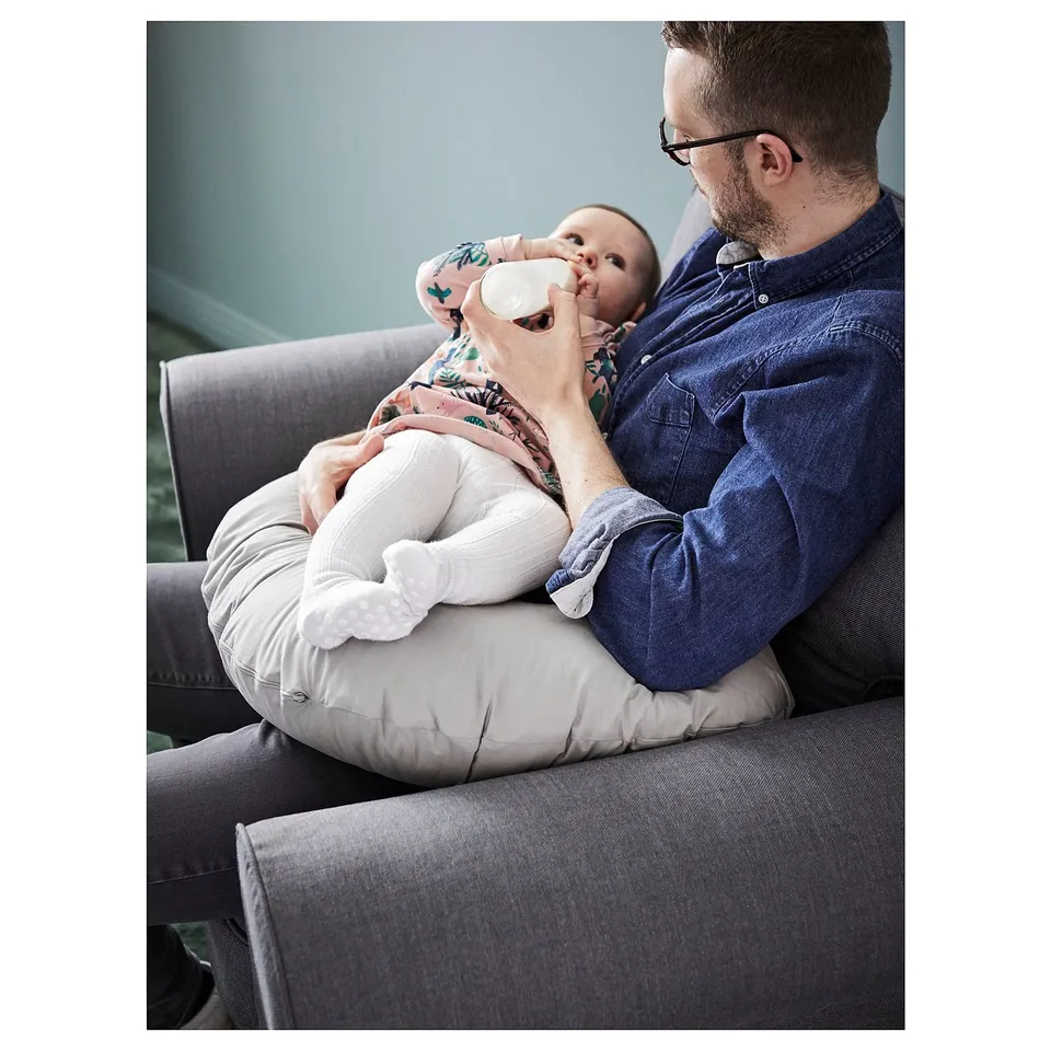 A person holds a baby with a nursing pillow. The pillow is grey, soft and round, supporting the babys feed.