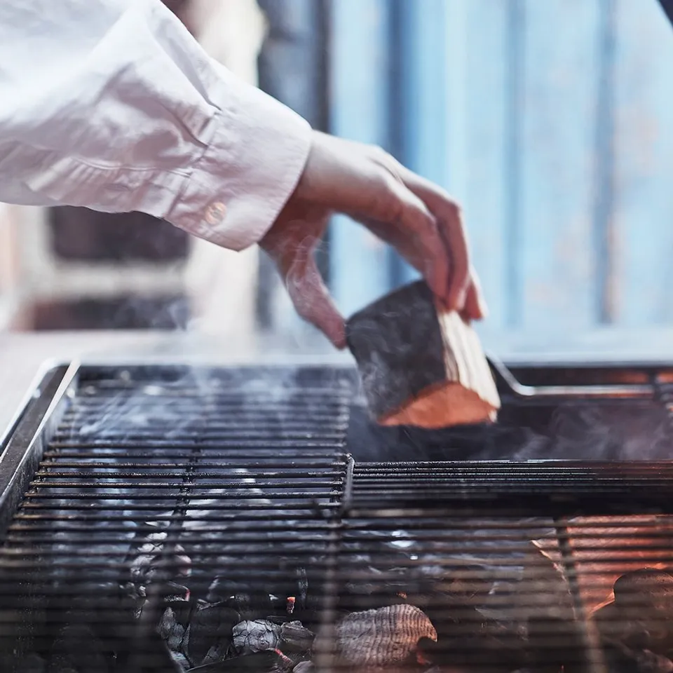 A person grills on a grilskär charcoal grill, using tongs. The grill has a stainless steel grate and visible flames.