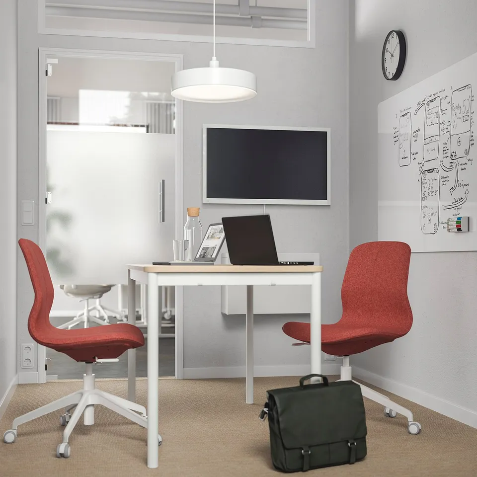 Minimalist office with two red LÅNGFJÄLL chairs, a white desk, and a tv. A black bag rests beneath the desk. The room is well-lit and organised.