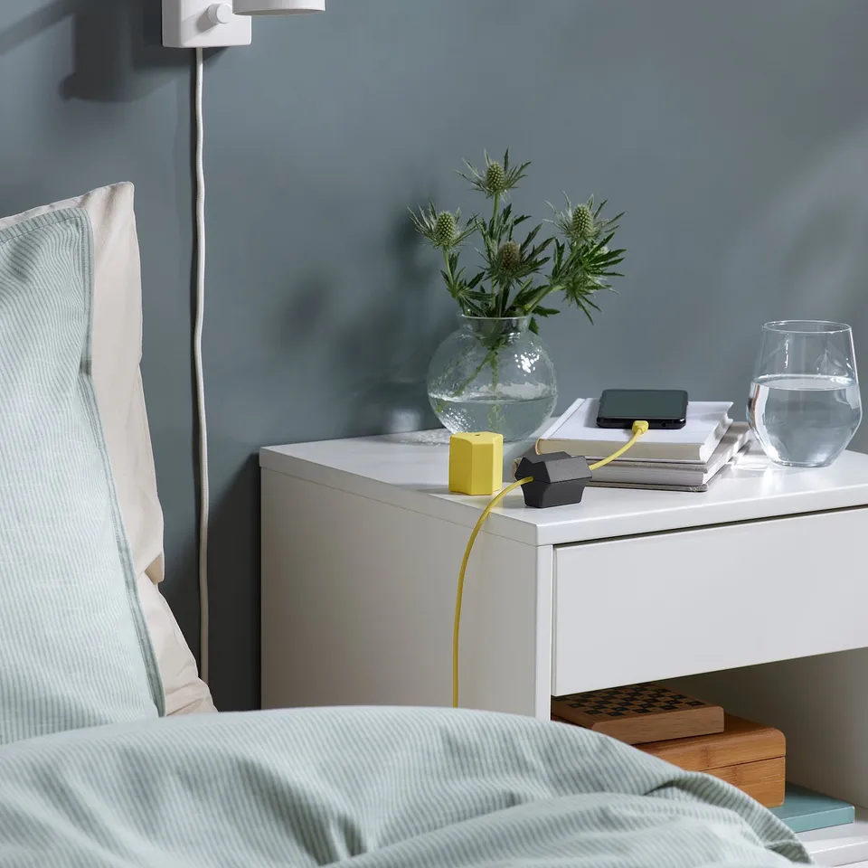 A bedside scene with a yellow HAVSKÅL usb anchor securing a cable on a white bedside table.