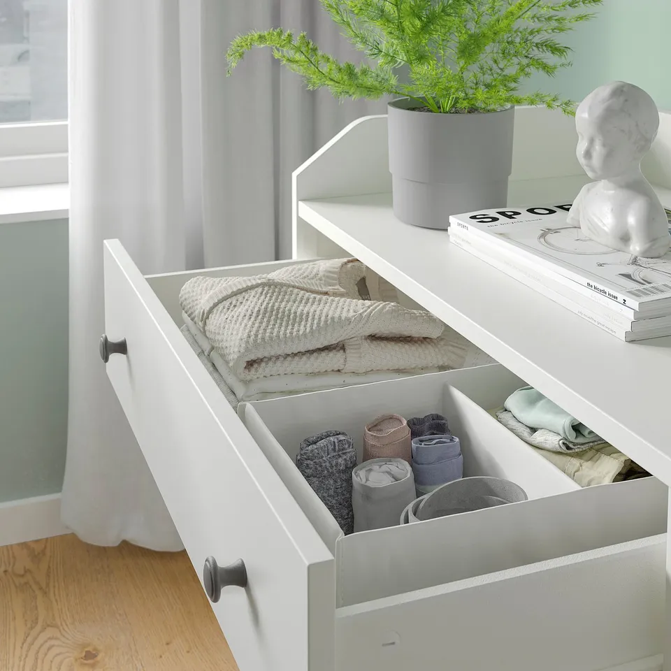 Open white HAUGA dresser with folded clothes, including sweaters and rolled socks, near a window with a plant and decorative bust.
