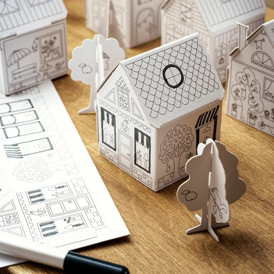 Image displays MÅLA paper craft houses, unpainted, black outlines, various shapes, on wooden table, with instructions.