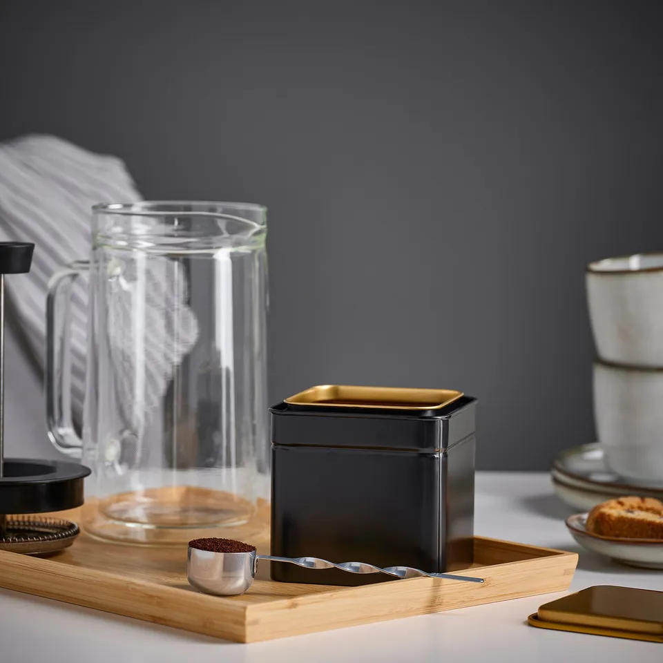 A wooden tray with a metal coffee scoop, a black container, a glass pitcher, and mugs. The scoop has a long handle and is filled with coffee grounds.