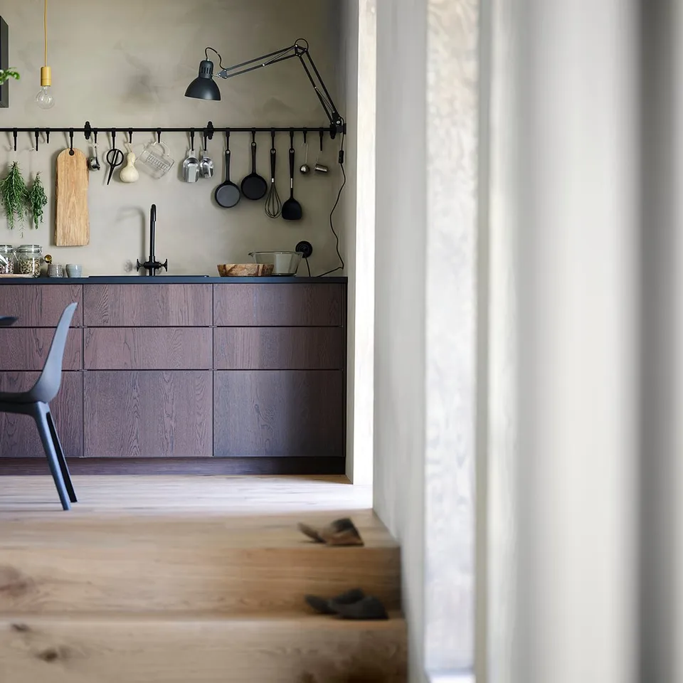 Modern kitchen with dark brown wooden cabinets, black chairs, hanging utensils, and green plants.