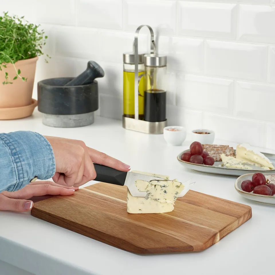 Person slices cheese on wooden board, plates, mortar nearby. Alternatively: cheese sliced on wooden board with food plates and mortar.