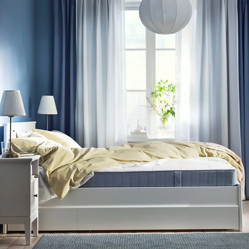 Modern bedroom with blue curtains, white bed, yellow blanket, lamps, and plant by window.