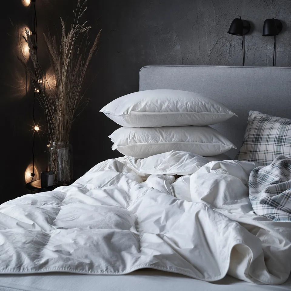Unmade bed with white FJÄLLBRÄCKA duvet; white pillows, plaid pillow case, dark wall with dried plants, lamps.