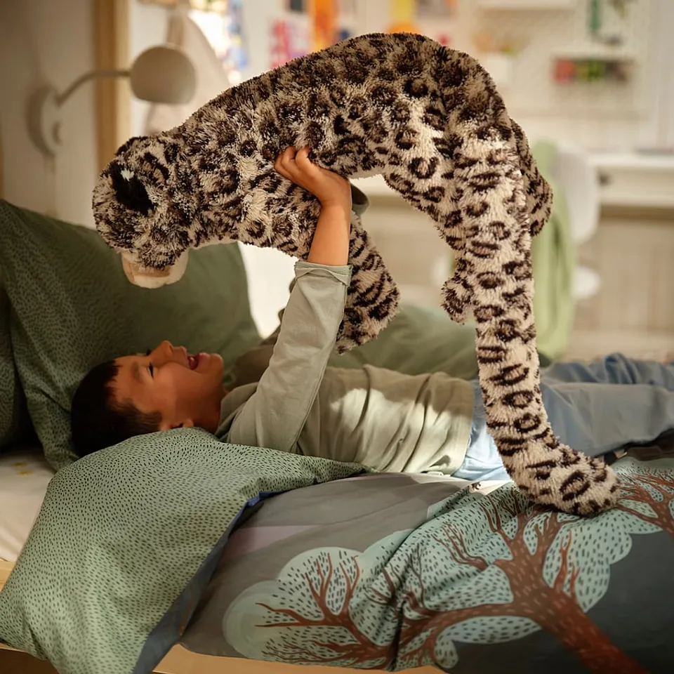 Child lies on rare double-sided duvet, showing himalayan scene and green speckles, raising plush snow leopard toy above.