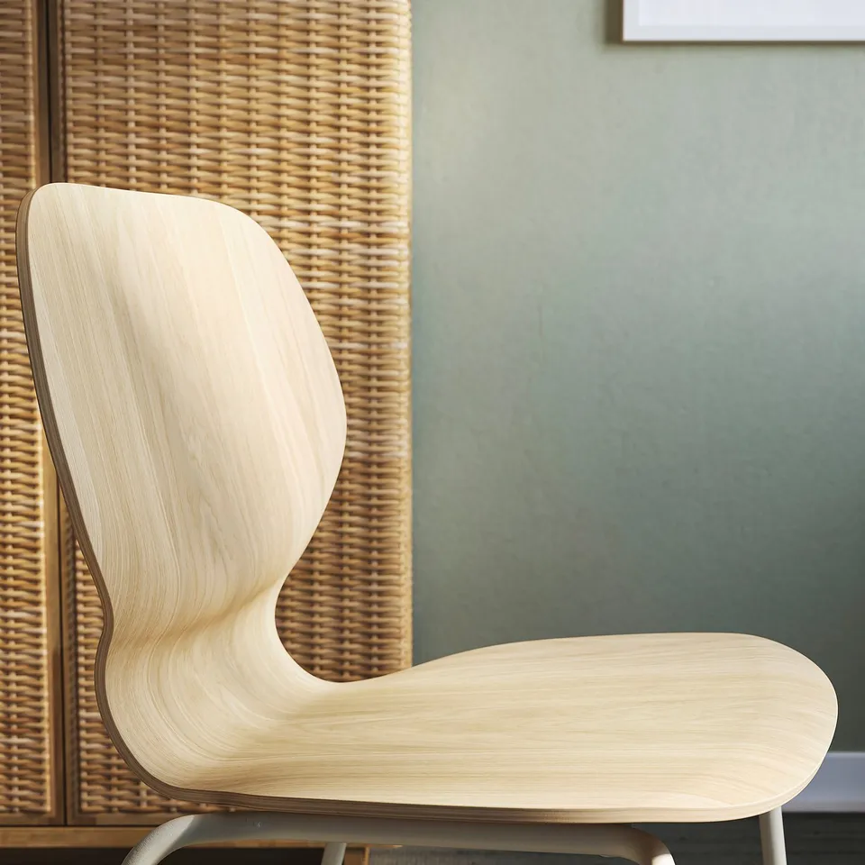 Modern wooden chair with smooth, curved edges and light finish. Positioned near a woven divider and green wall.