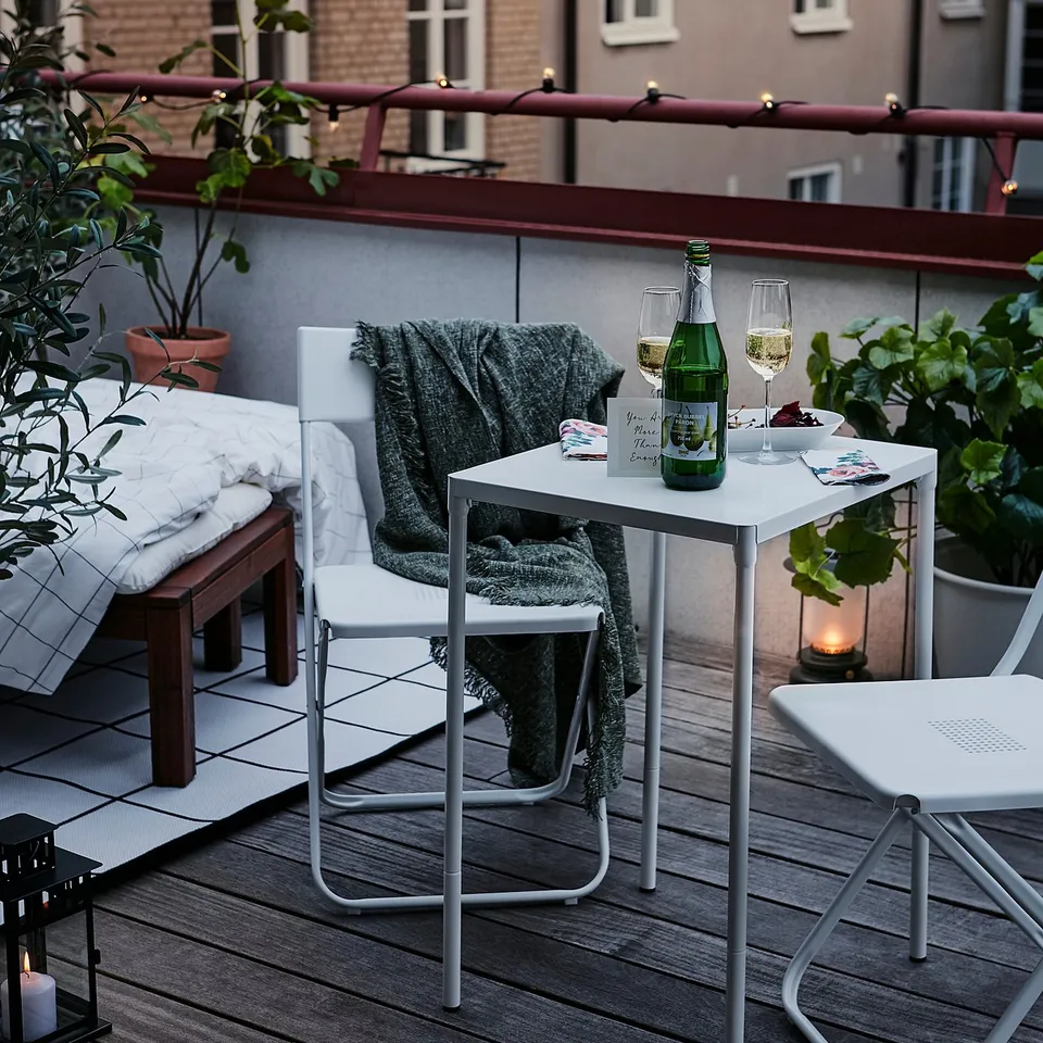 Modern balcony with white metal table, drink bottle, wine glass, blanket, lantern, and potted plants.