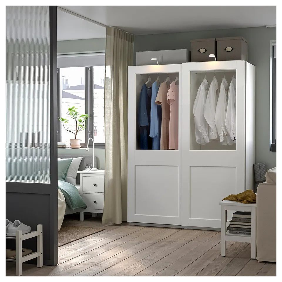 Room with a white PAX wardrobe. Clothes hang inside, visible through glass doors. A bedroom setting with a bed and shoe rack nearby.