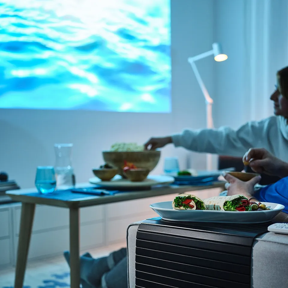 Person eating wrap on sofa with RÖDEBY bamboo tray. Blue-themed room, wall screen displaying water.