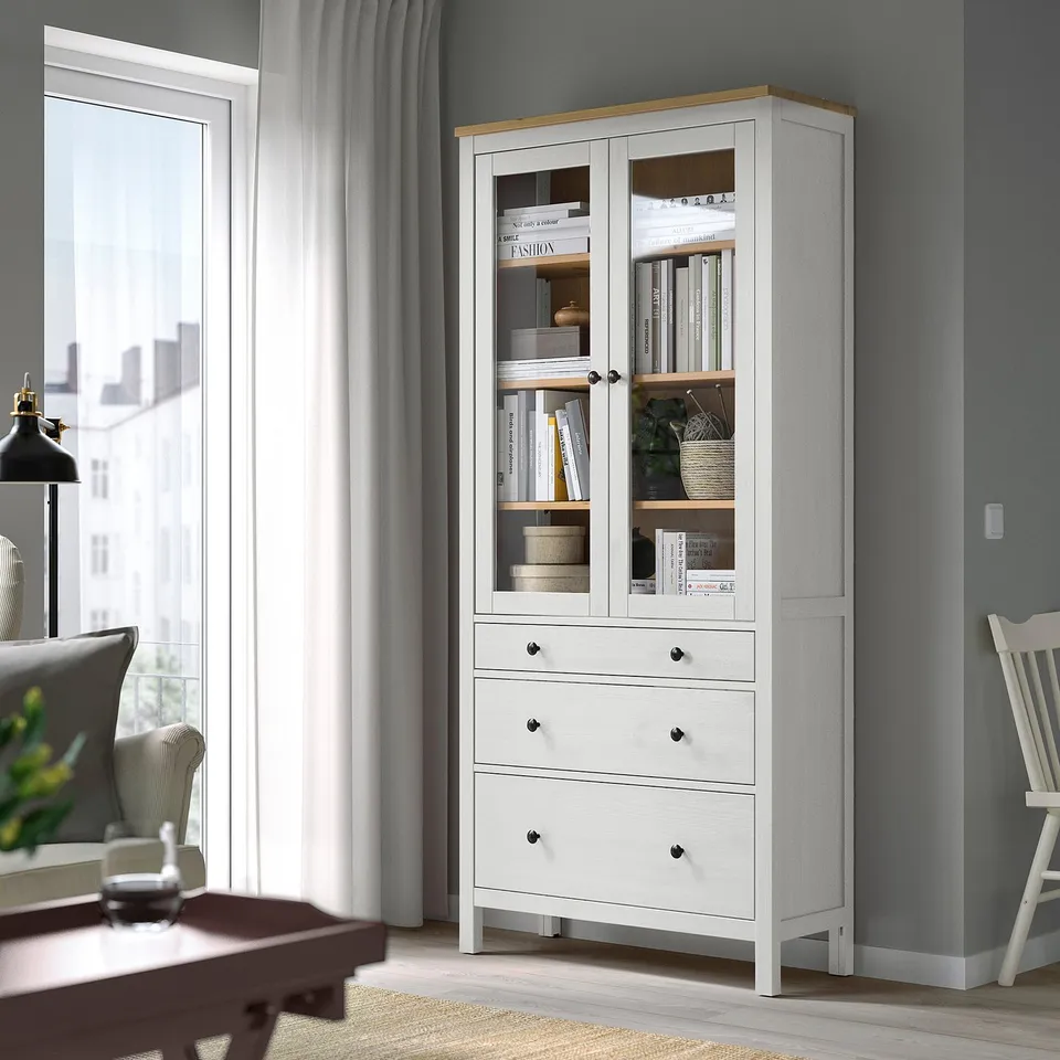 Wooden HEMNES cabinet with glass doors, storing books and items, between window and chair.