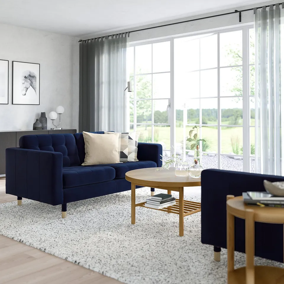 Modern room with large windows, blue landkrona sofa, wooden coffee table, grey rug, and side tables. Sofas size complements the rooms layout.