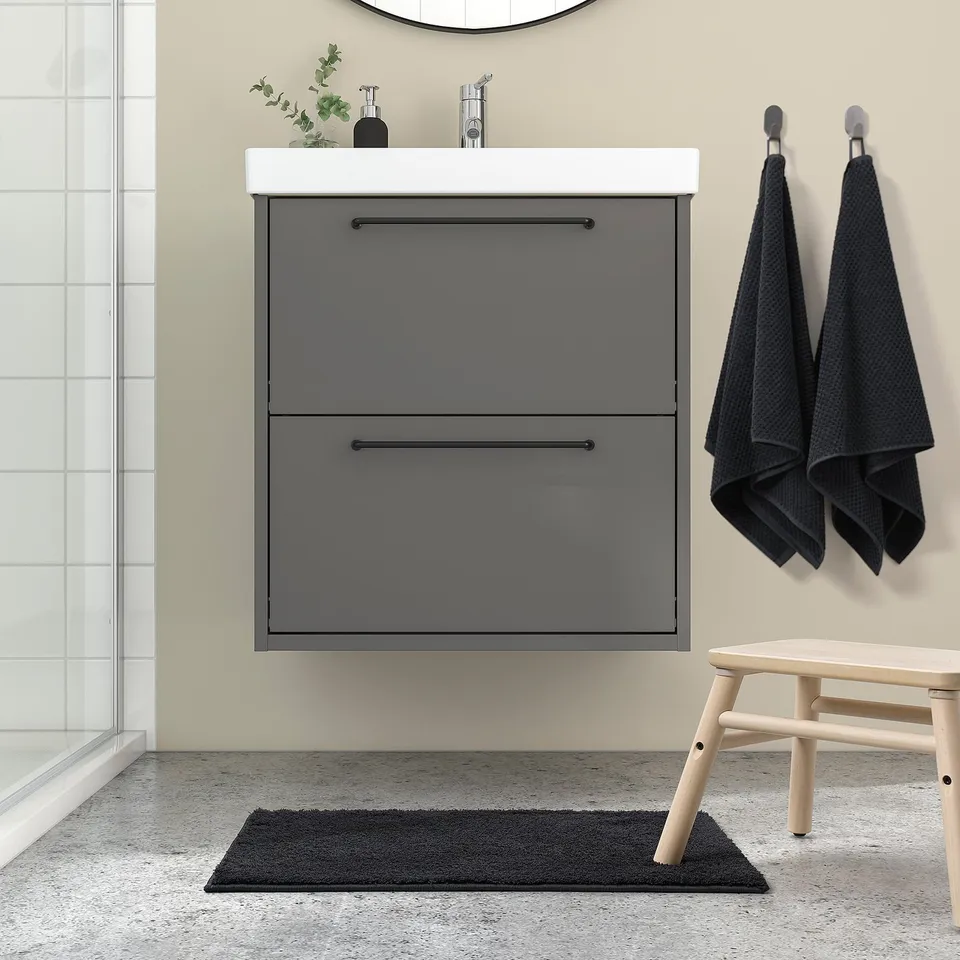 Modern bathroom with grey BACKMÅRA mat, wooden stool, hanging towels, and sleek vanity.