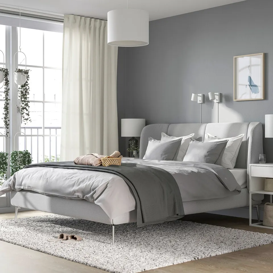Modern grey bed with curved headboard, plush pillows, large window, plant, cosy vibe.