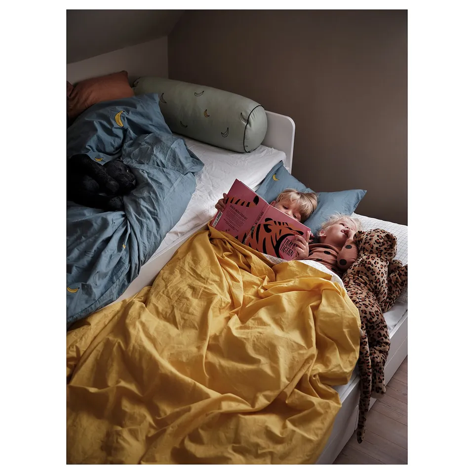 Two children read in bed with yellow blanket, blue pillow, stuffed animals, and open book.