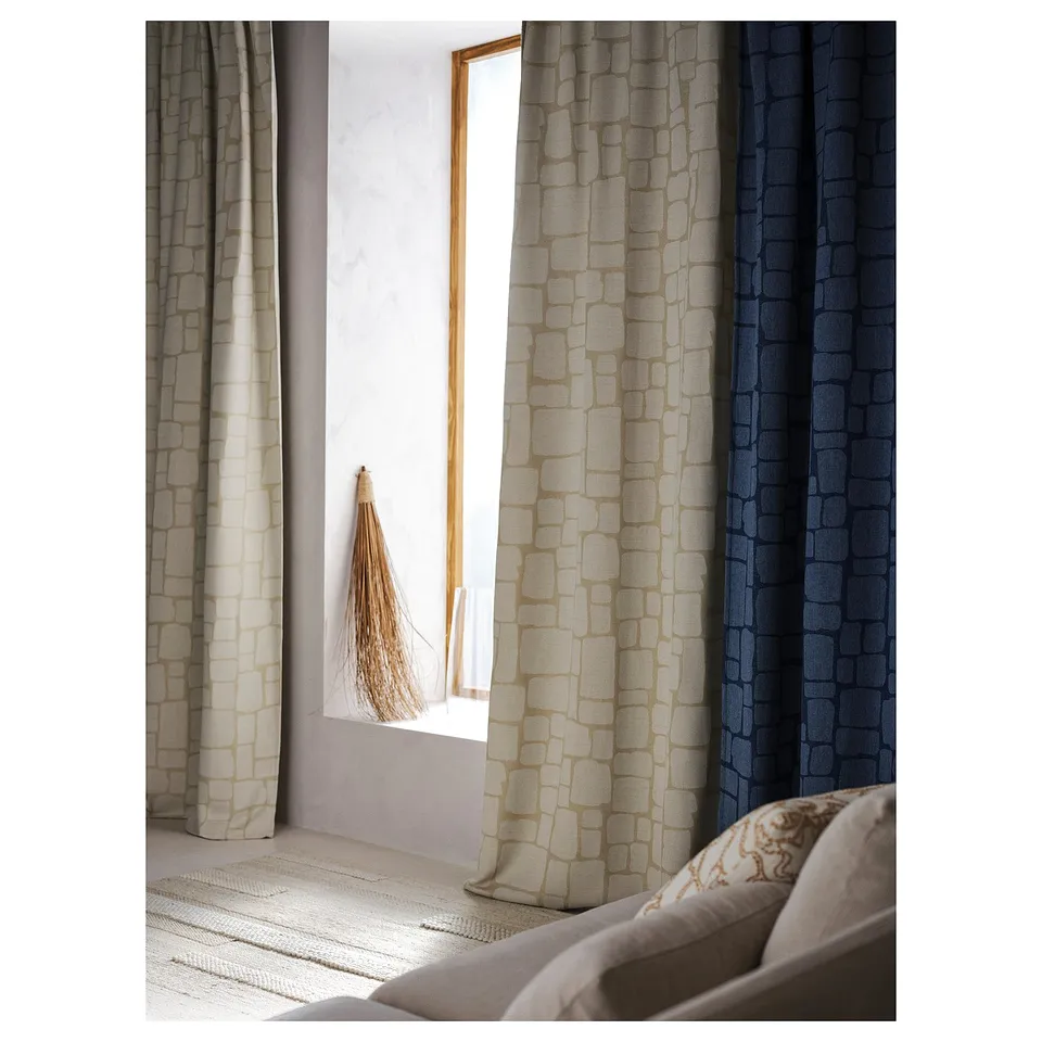 Three draped curtains in beige, beige-brown, and navy with pattern, beside a window, near a bed with a pillow.