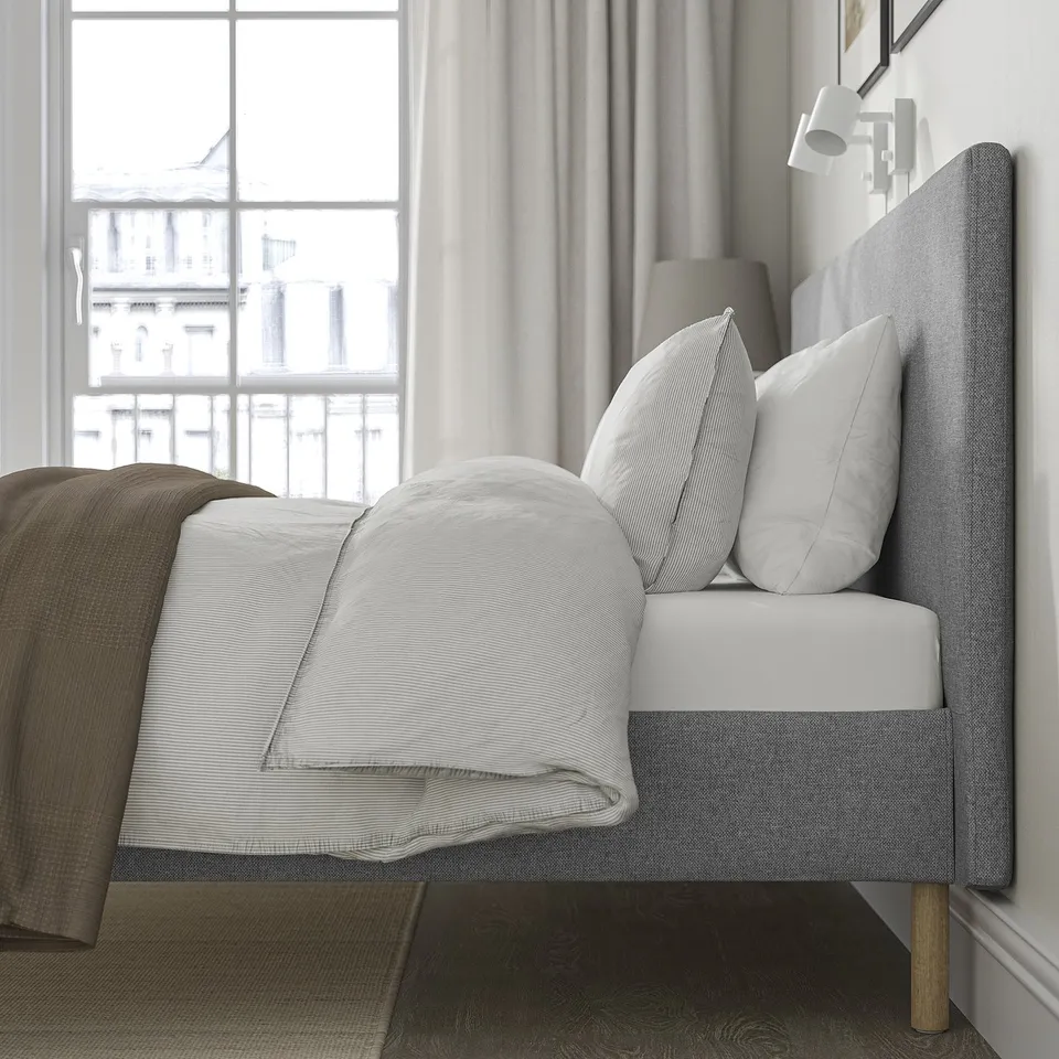 Bed with grey upholstered headboard, white pillows, and blanket. Padded headboard for comfort. Clean, minimalist design. Large window with curtains.