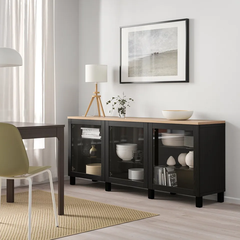 Black-brown SINDVIK sideboard with glass doors, white dishes, framed wall, lamp, green chair beside.