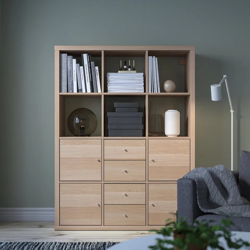 KALLAX light wood bookshelf with storage. Holds books, decor. Tall, detailed, near grey couch. Has shelves, drawers.