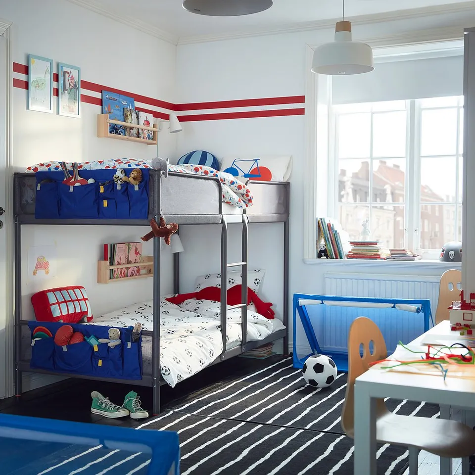 Kids bedroom with blue & red bunk beds, colourful bedding, white walls, striped rug, blue storage, books, toys, and a large window.