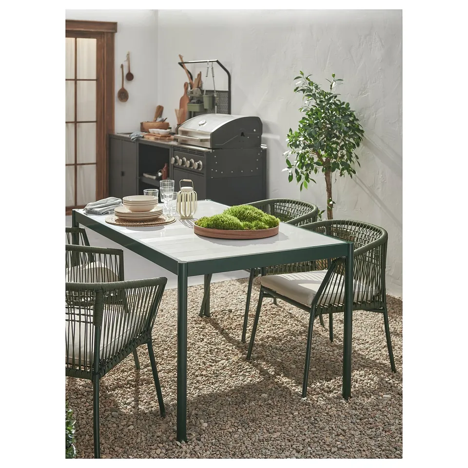 Modern outdoor dining set with rectangular table, four chairs, and decorative plants. Dark green frames and beige cushions.