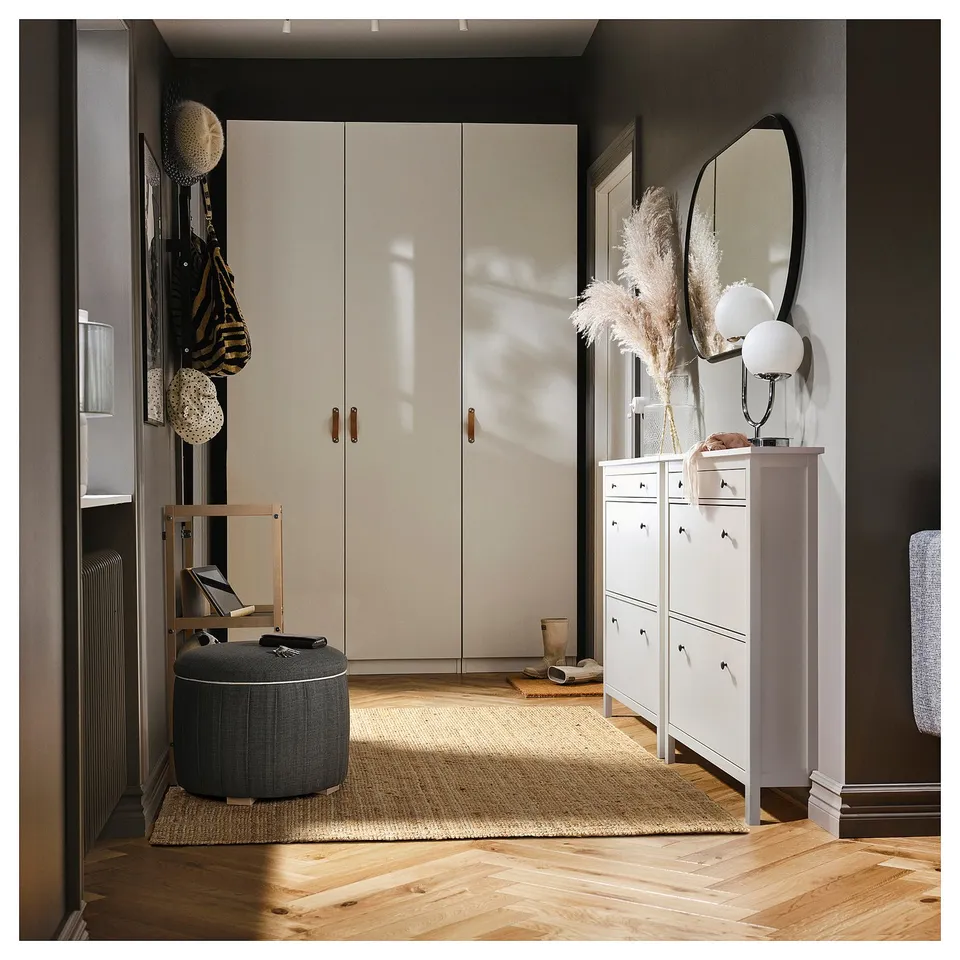 Modern entryway with white furniture: tall closets, shoe cabinet, round ottoman, wooden floors, and mirror.