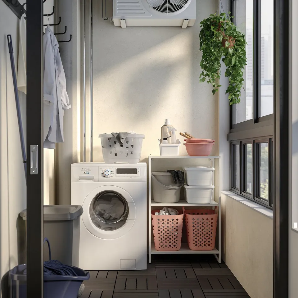 Compact laundry room with washing machine, pink/white baskets, plant, cleaning supplies, coat rack with shirt, dark wood floors.
