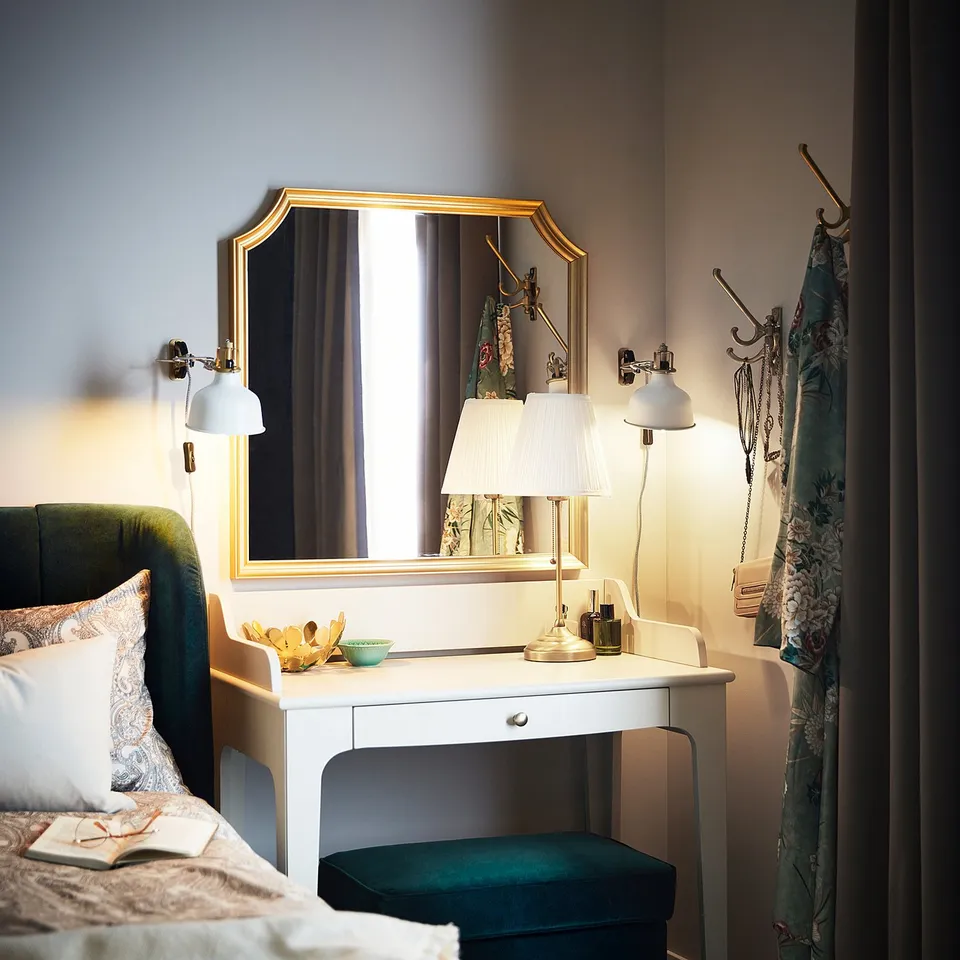 Bedroom with green bed, white desk, gold-framed mirror, lamp, and blue stool. Curtains and jewellery on the wall.