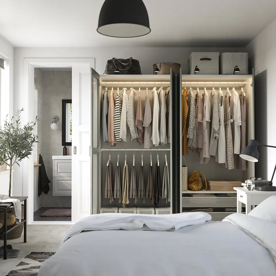 Modern bedroom with open PAX wardrobe displaying clothes on hangers and shelves. Mirror doors and adjustable interior organisers.