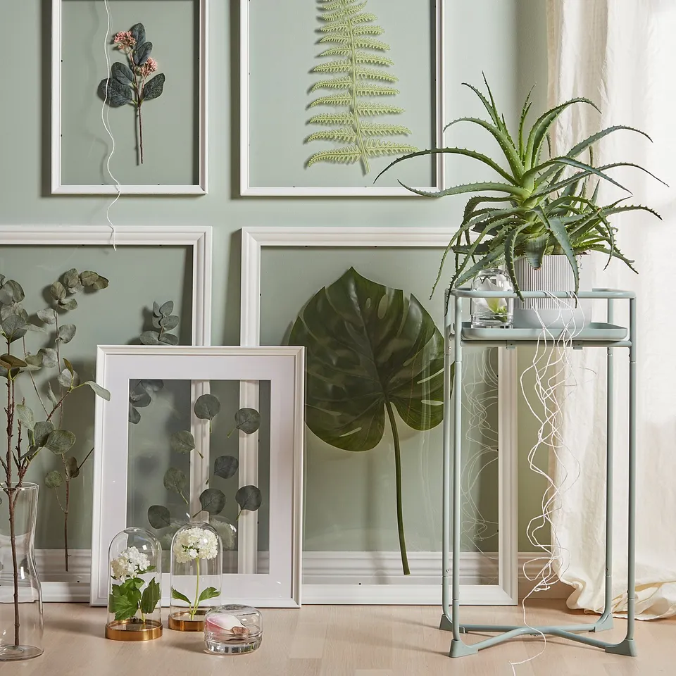 A cosy corner with framed floral art and a potted plant on a metal stand against a light green wall.