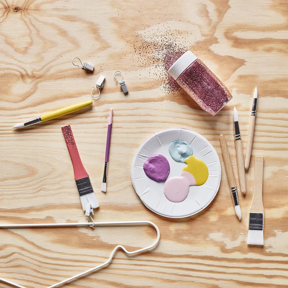 A creative workspace with MÅLA tools: colourful paint palettes, brushes, and glitter jar on a wooden table.