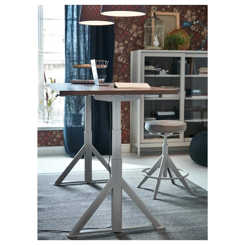 Home office setup with adjustable desk and stool, laptop, bookshelf, and pendant lights.