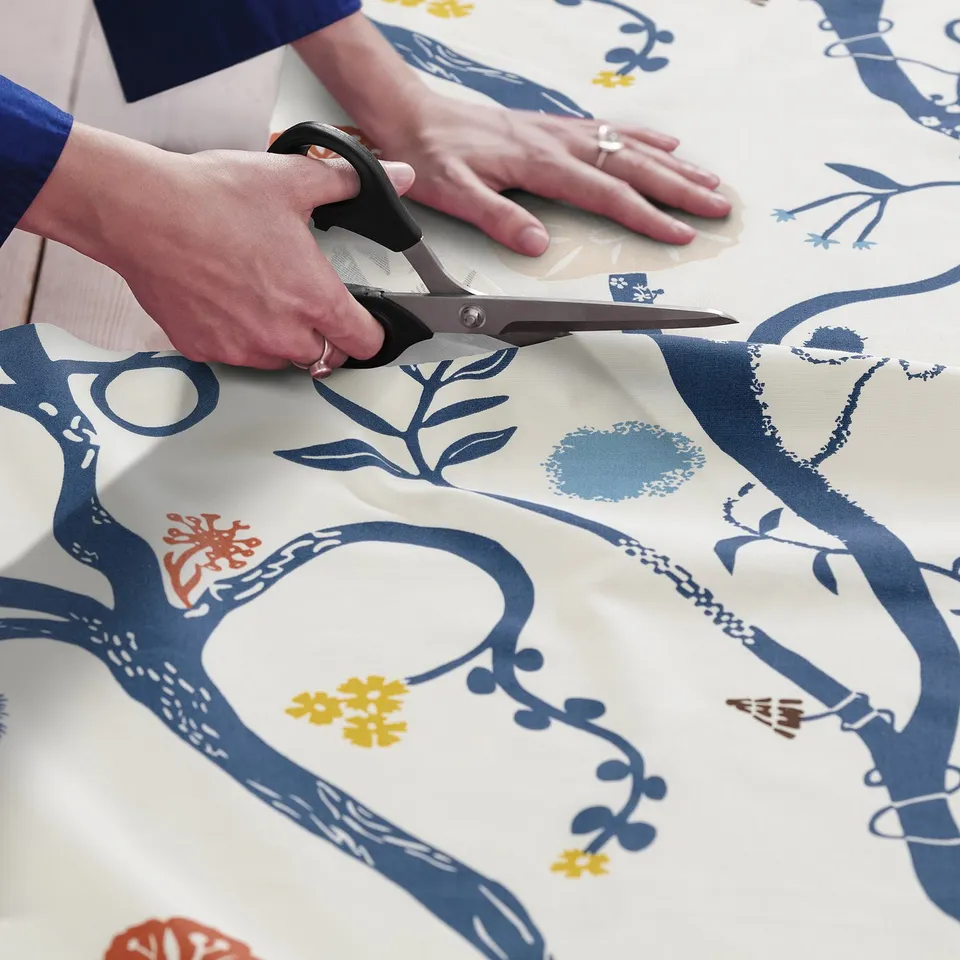 A person cuts REVFINGERÖRT fabric with scissors. The fabric a whimsical meadow design with flowers and winding patterns, ideal for sewing projects.
