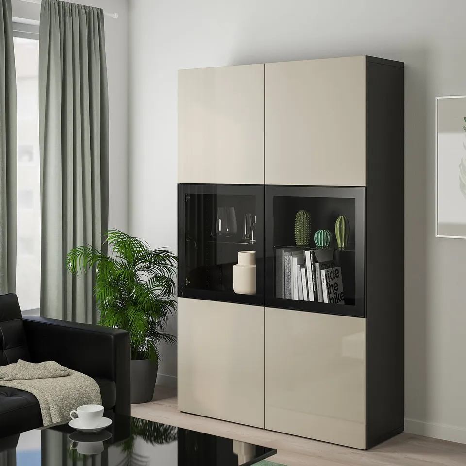 Large white and black BESTÅ cabinet, glass doors, showing decor and books, next to potted plant, black chair, and cup on table.