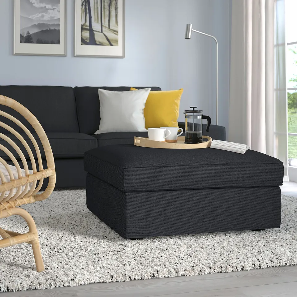 Modern living room with grey ottoman, black couch, yellow pillow, wooden floor, wicker chair, and wall decor.
