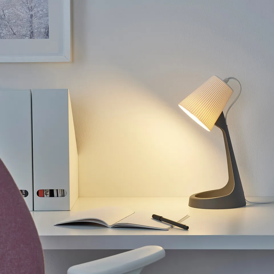 SVALLET lamp on a desk with books and a chair. Black lamp with a ribbed white shade, lit.