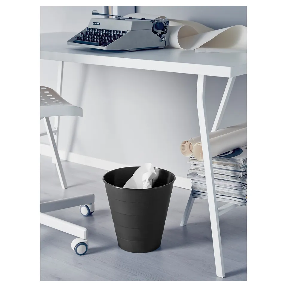 Black rubbish bin under a white desk with casters, holding crumpled white paper.