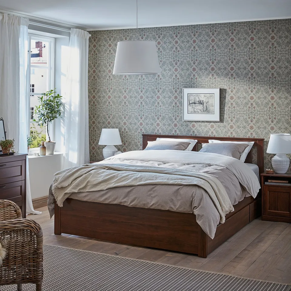 Bedroom with dark wooden songeasnd bed, beige bedding, patterned wallpaper, white curtains, table lamps, wicker chair, plants, and hanging light.