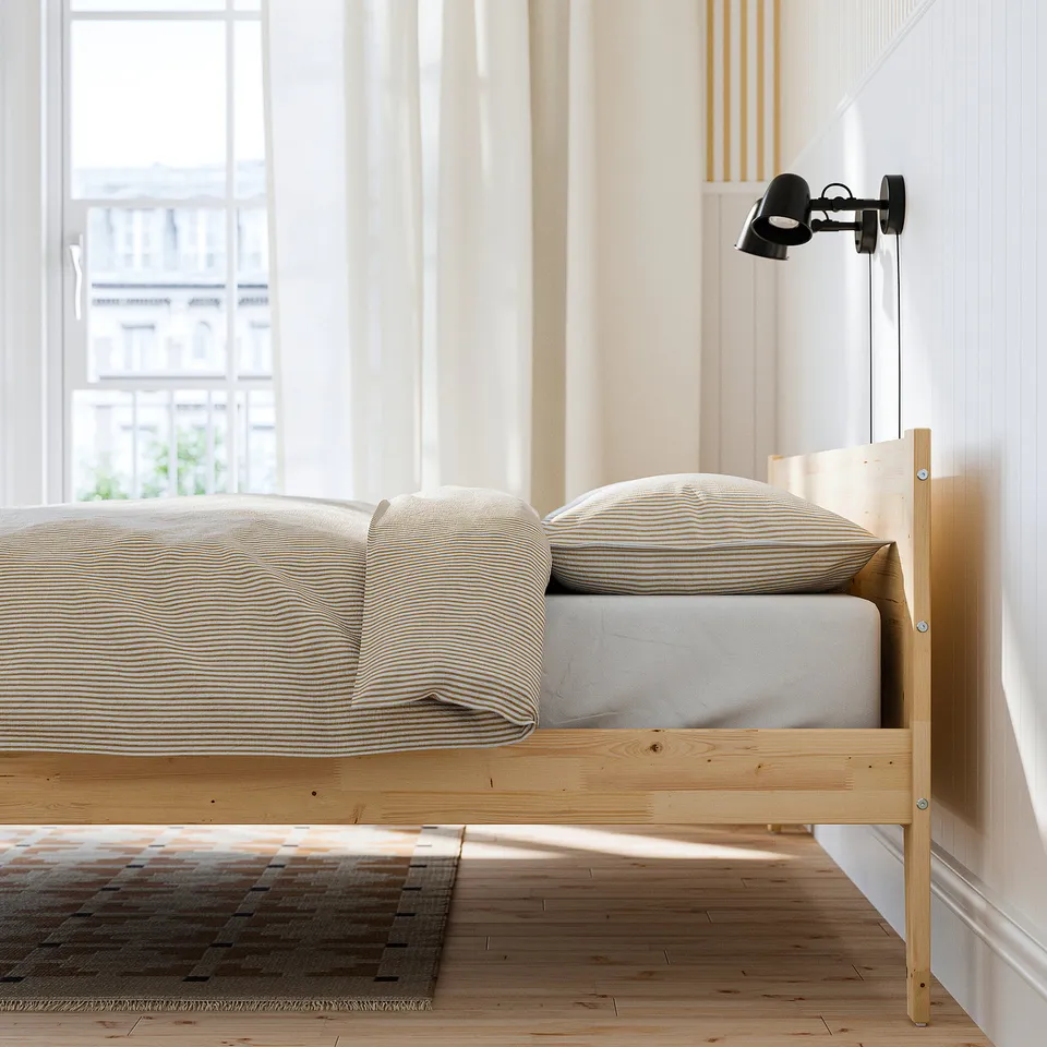 Bedroom with taxasklint wooden bed frame, striped bedding, wall-mounted black lamp, and large window with sheer curtains.