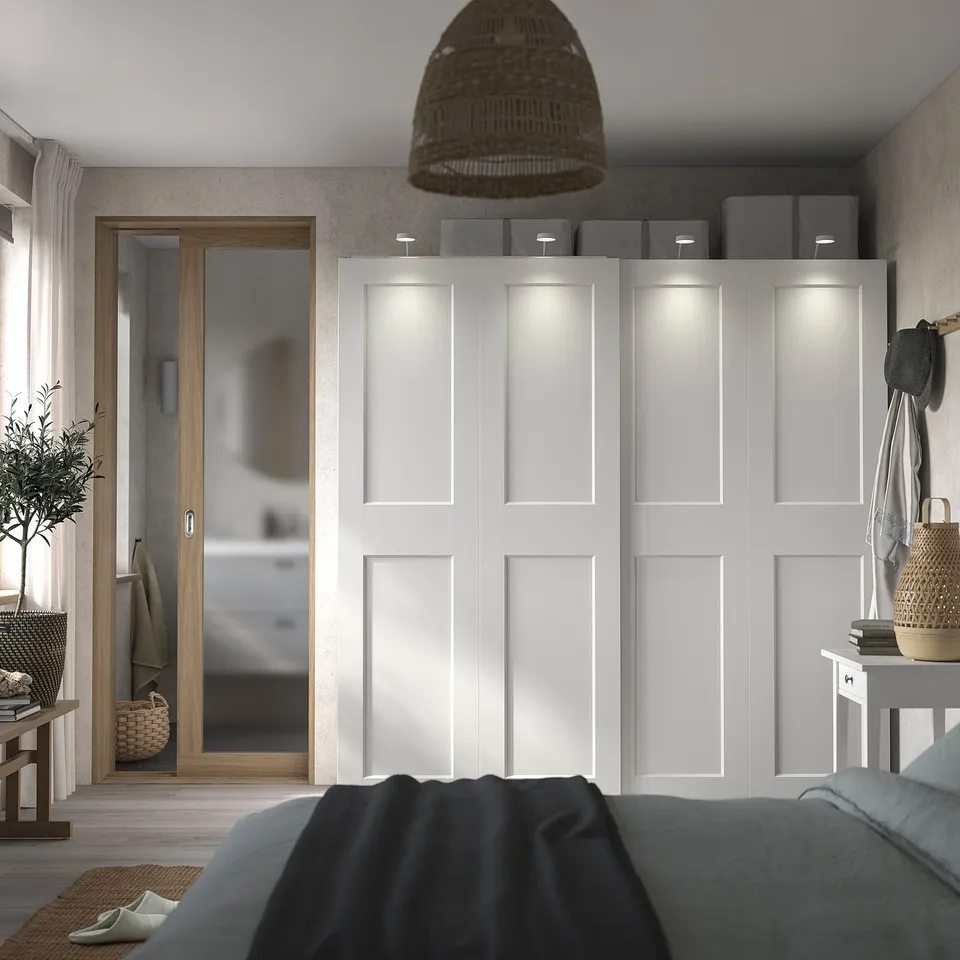 Modern bedroom with grey PAX wardrobe, sliding doors, and top shelves holding bins. A wooden door leads to another room.