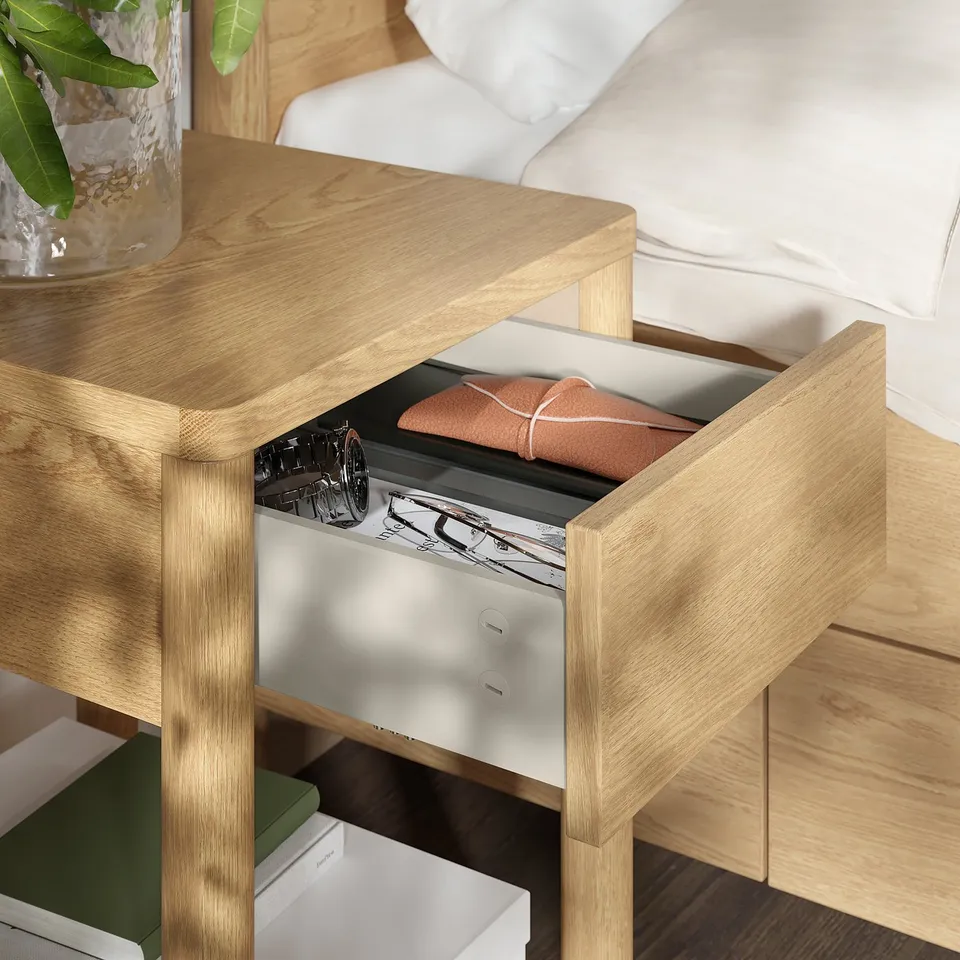 Open wooden bedside drawer reveals neatly folded orange cloth, beside black-and-white art book, atop light wood bedside table next to bed.