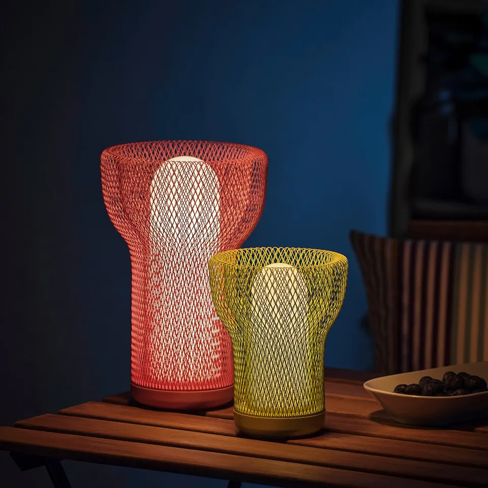 Two SOLVINDEN lamps; red & yellow, on table. Mesh design, indoor/outdoor use. Red is taller, both modern & decorative. Bowl of fruit beside.