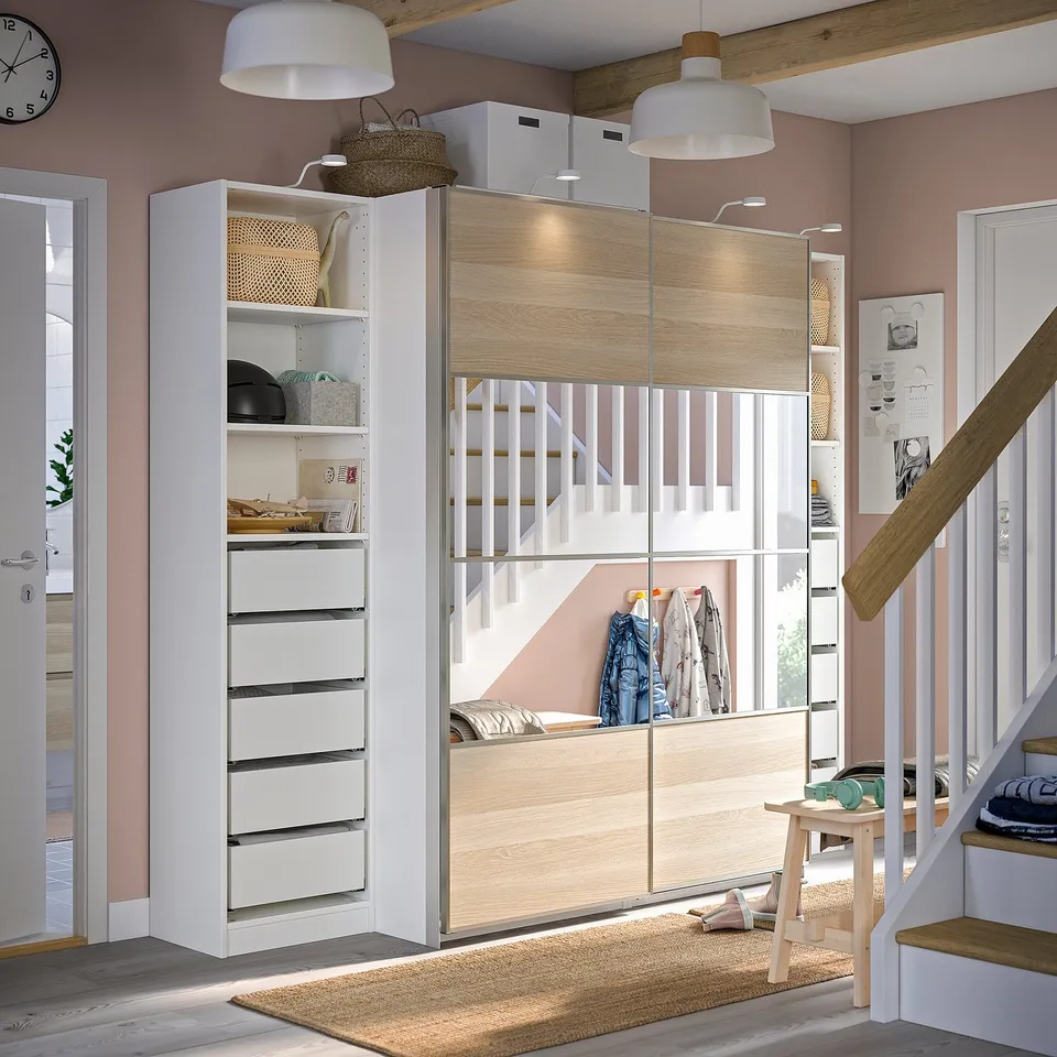 A room with a white and light wood PAX wardrobe. It has drawers and sliding doors with built-in lighting. Theres a stairway on the right.