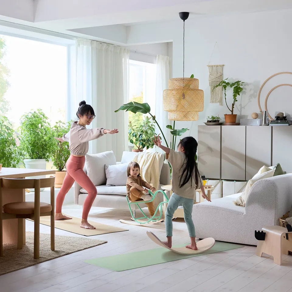 Family playing with greejimojs balance board near couch and window, greenery in background.