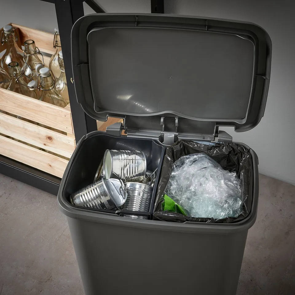 Black dual-compartment recycling bin with one open lid, revealing cans in one section and plastic waste in the other.