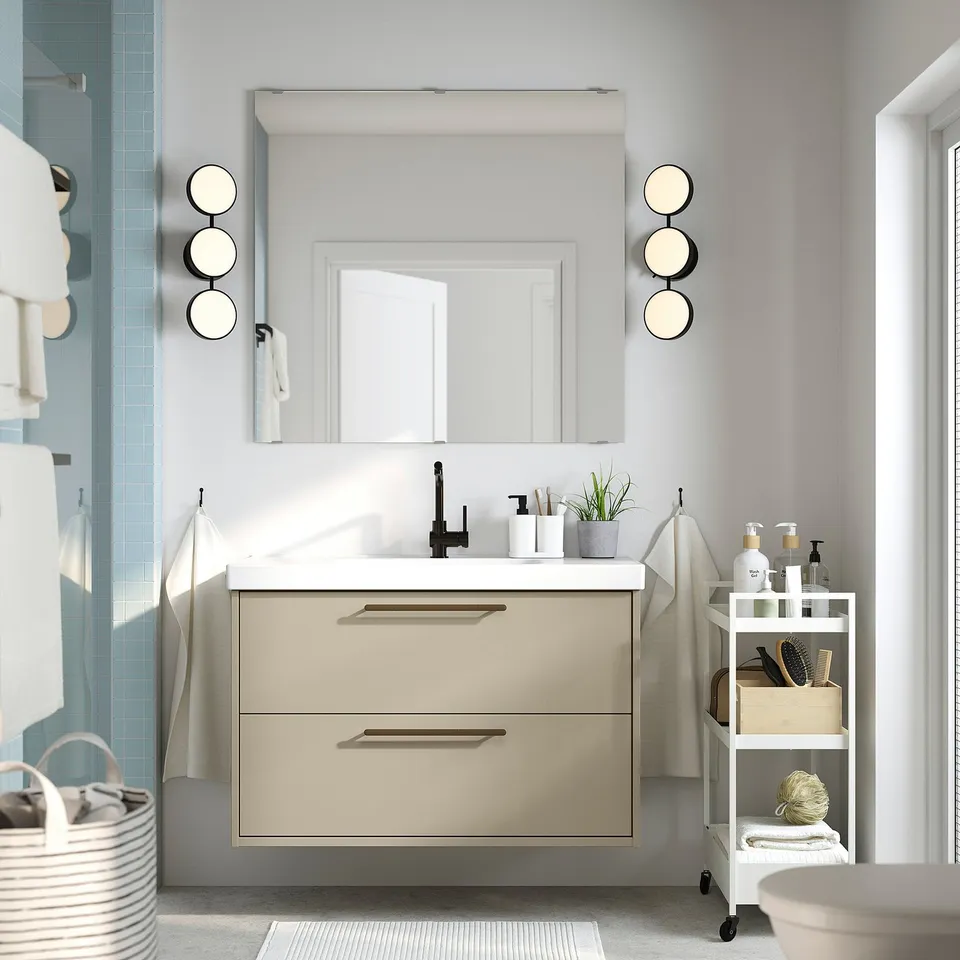 Modern bathroom with HAVBÄCK vanity, mirror, sink, black fixtures. Wall-mounted, deep drawers. White cart, towels, plants.